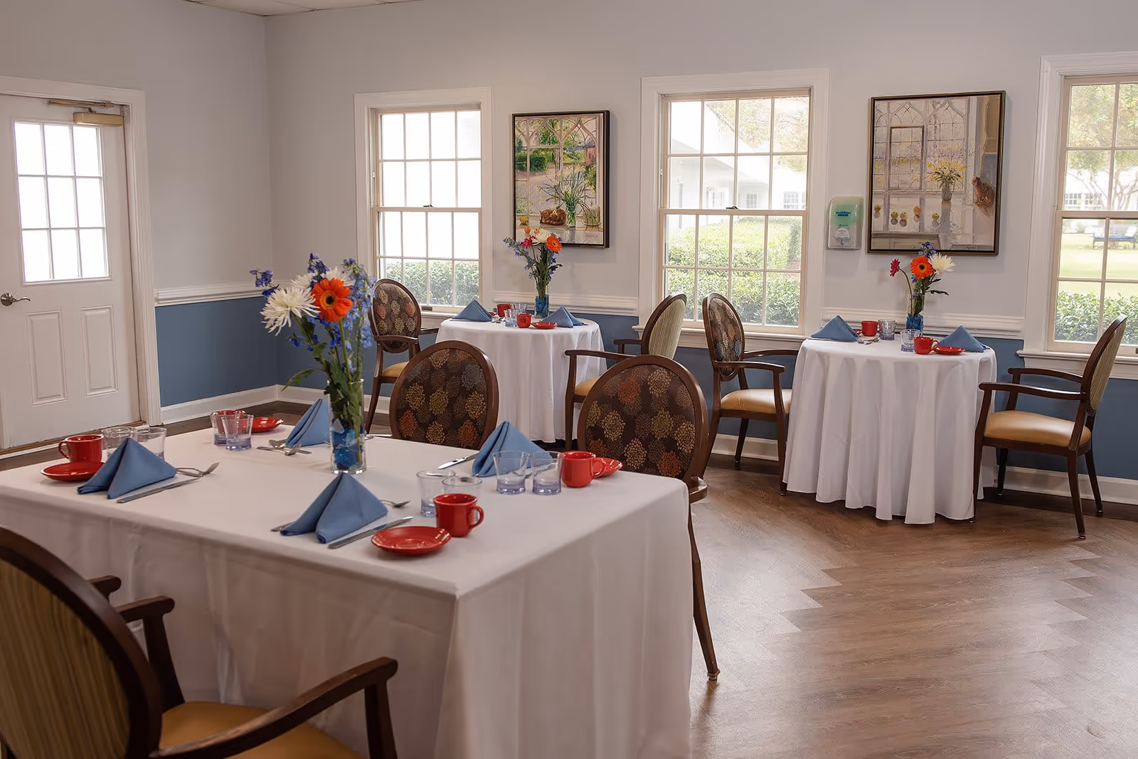 A dining room with tables covered in white tablecloths, each set with blue napkins, red cups and plates, and glassware. The tables have floral centerpieces with colorful flowers. The room has large windows letting in natural light, framed artwork on the walls, and wooden chairs with patterned upholstery.