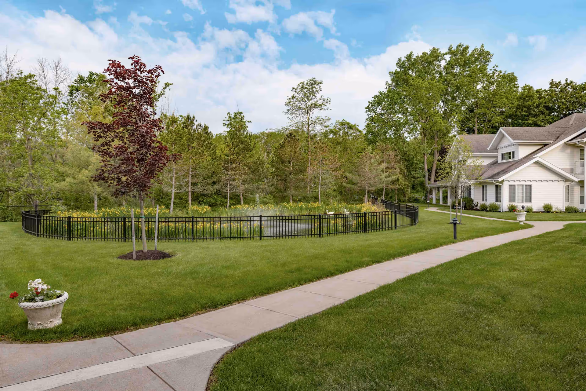 A well-maintained outdoor area at Holiday Maple Downs featuring a curved concrete walkway, green lawn, a small tree with red leaves, a fenced pond with yellow flowers, and a white building with a porch surrounded by trees under a partly cloudy sky.