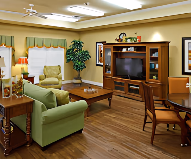 A cozy living room with a green sofa, an armchair, a wooden coffee table, and a wooden entertainment center with a TV. There is a round dining table with chairs on the right side, a potted plant in the corner, and framed artwork on the walls. The room has wooden flooring and beige walls with green valances on the windows.