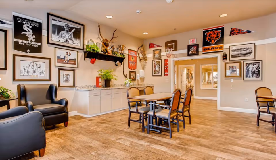 A cozy common area in a senior living facility decorated with sports memorabilia including framed photos, banners, and trophies. The room features a wooden floor, a table with six chairs, two black armchairs, and a countertop with cabinets underneath. The walls are adorned with Chicago White Sox and Chicago Bears decorations.