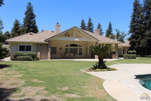 Single-story house with a covered patio area furnished with a table and chairs, surrounded by a well-maintained lawn, trees, and a swimming pool in the foreground under a clear blue sky.