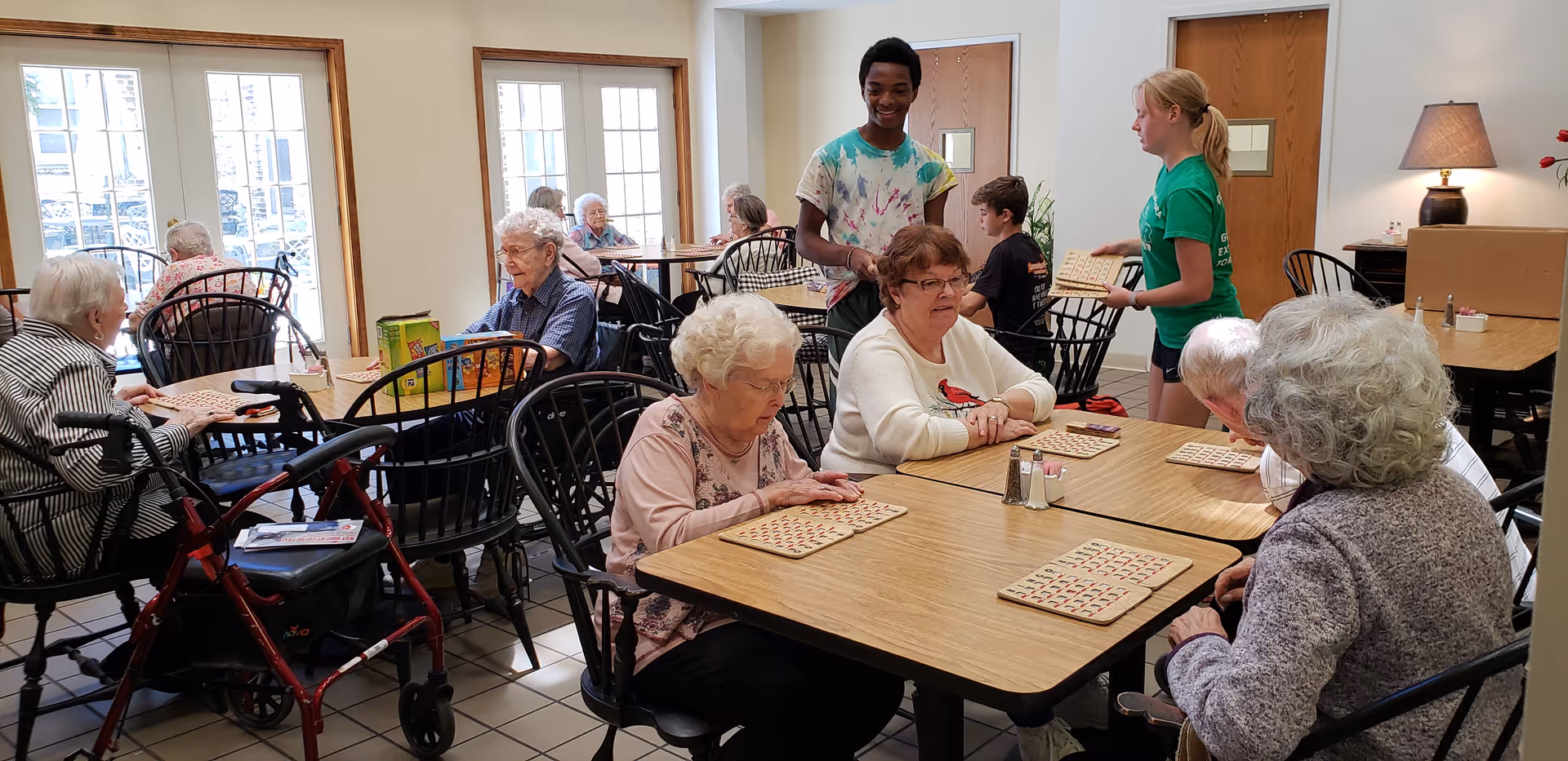 Seniors seated at tables in a bright common room playing bingo while two younger volunteers assist.