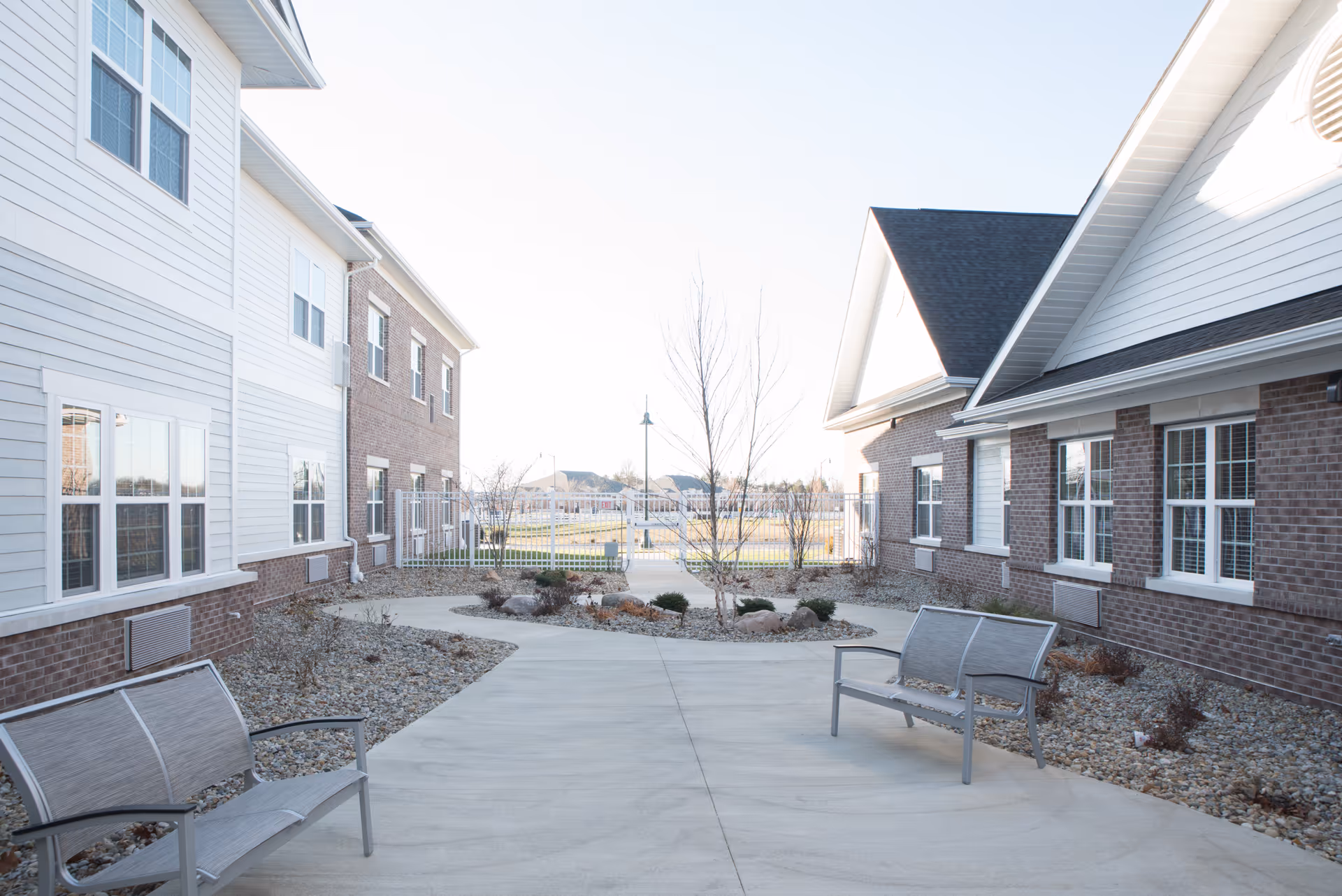 Outdoor courtyard area between two buildings with benches, small trees, and landscaping with rocks and shrubs under a clear sky.