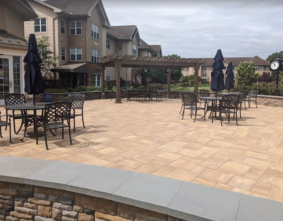 Outdoor patio courtyard with metal tables and chairs, closed umbrellas and a wooden pergola in front of a multi-story senior living building.