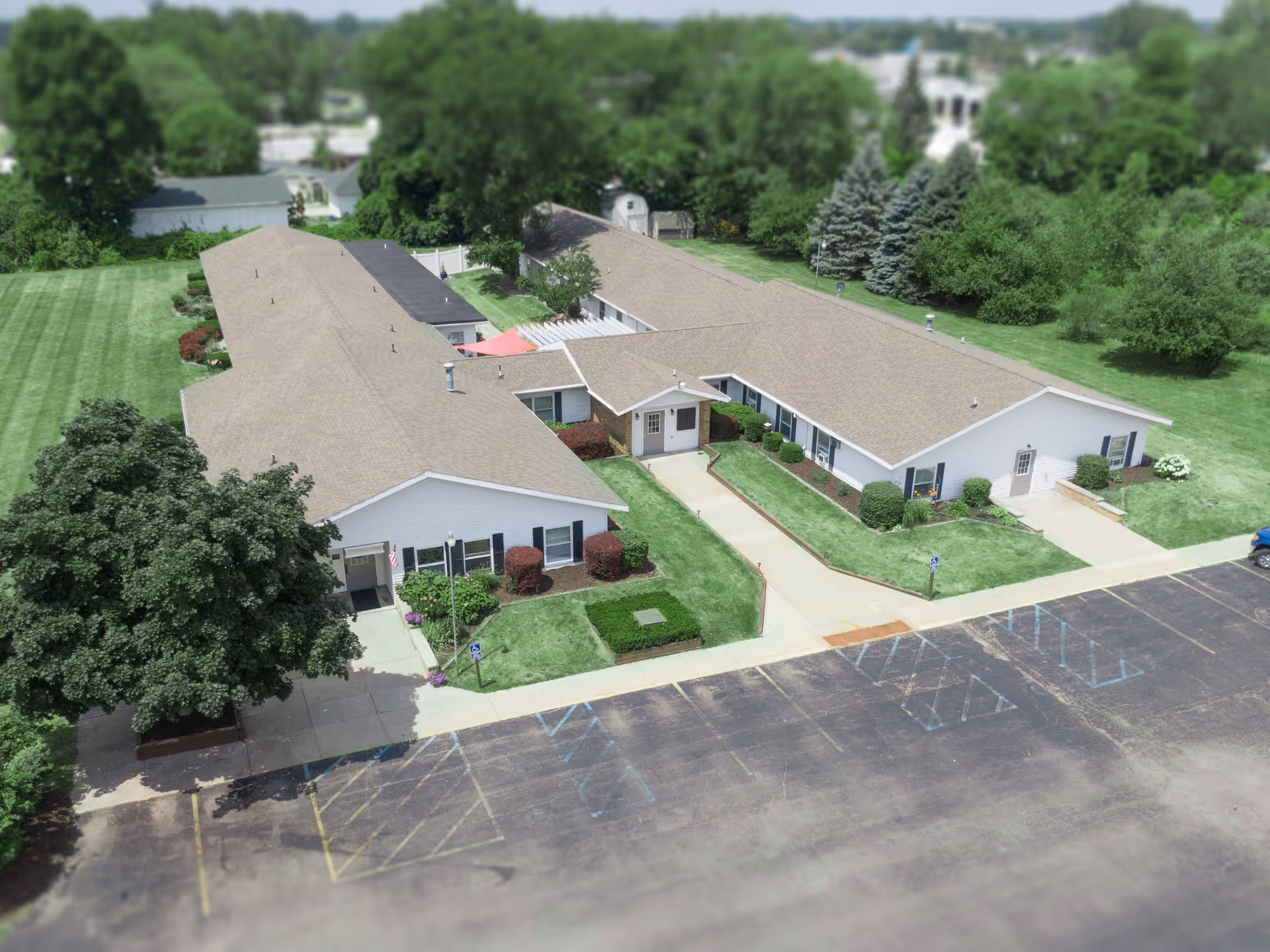 Aerial view of Divine Living Center of Mt Pleasant, a single-story senior living facility with beige roofs and white exterior walls. The building is surrounded by well-maintained green lawns, bushes, and trees. There is a parking lot with marked spaces in front of the building.