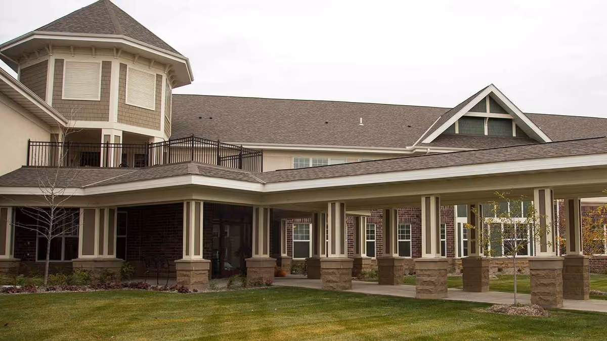 Exterior view of a senior living facility building with a covered walkway supported by columns, a turret-like structure, and well-maintained lawn and landscaping.