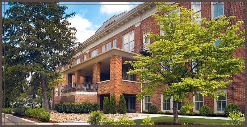 Exterior view of a multi-story brick building with large windows, a covered balcony supported by brick columns, surrounded by green trees and landscaping under a blue sky with some clouds.