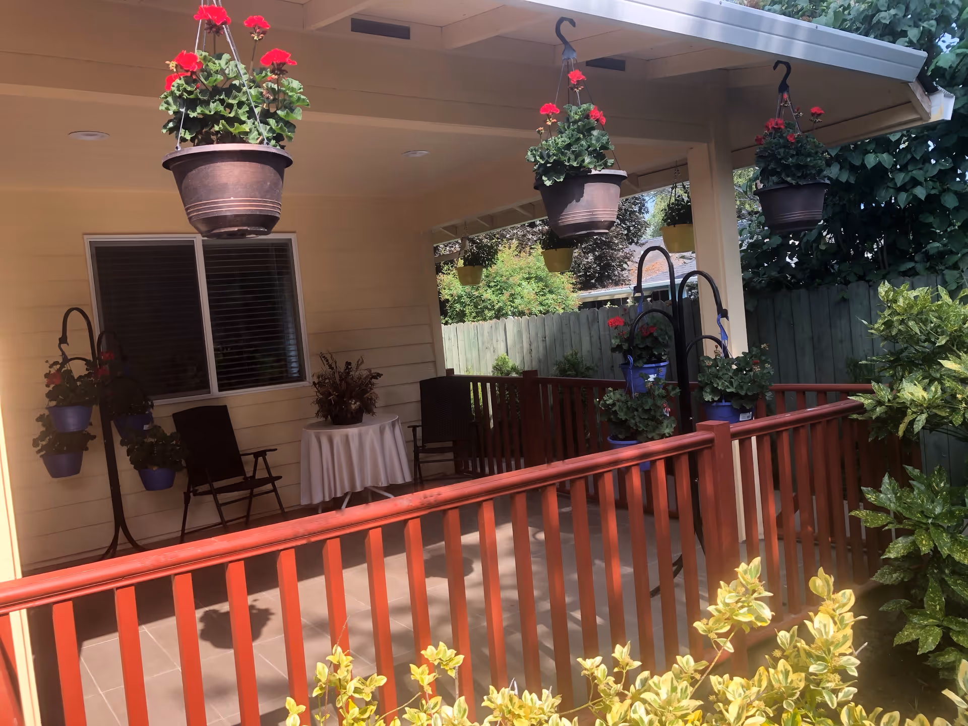 Covered outdoor patio area with hanging flower pots containing red flowers, a small round table with a white tablecloth, two black chairs, and a wooden railing. The patio is adjacent to a beige building with a window and surrounded by greenery and a wooden fence.
