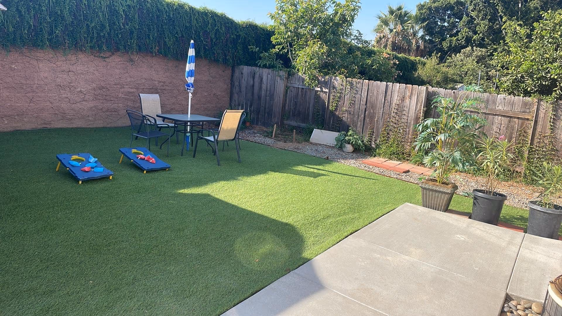 Outdoor area with artificial grass, a table with four chairs and a blue and white striped umbrella, two cornhole boards with bean bags, potted plants along a wooden fence, and a concrete patio in the foreground.