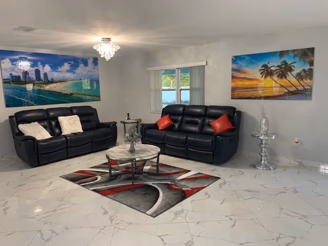 Bright living room with two black leather sofas, a round coffee table on a patterned rug, tropical wall art, and marble-look tile floors.