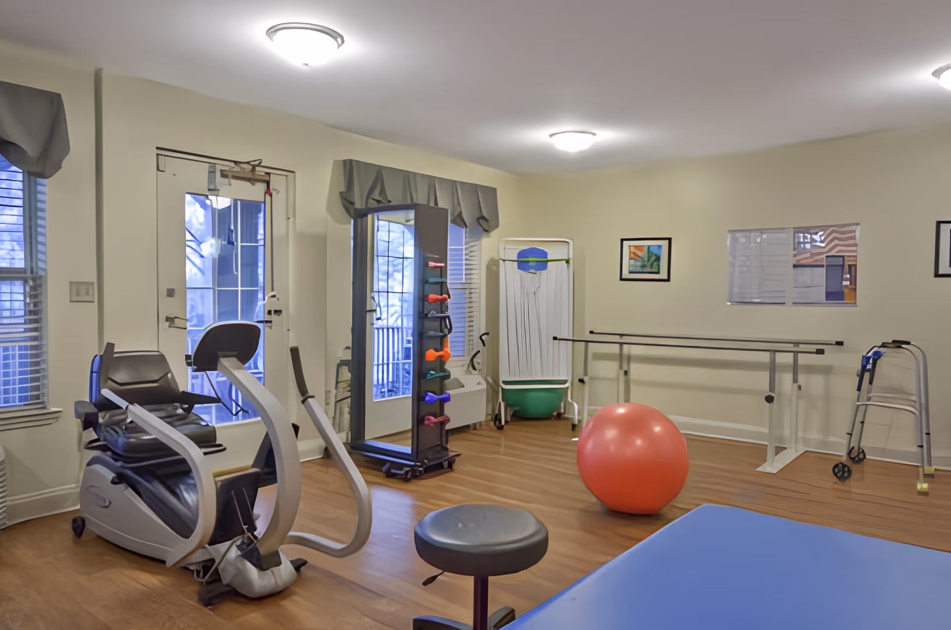 Small rehabilitation/exercise room with stationary exercise equipment, free weights, a large red therapy ball, parallel bars and a walker.