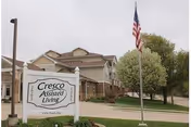Front exterior of Cresco Assisted Living building with a sign, American flag, and trees.