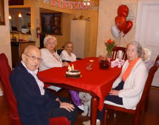 Four elderly people sitting around a red tablecloth-covered dining table in a cozy room decorated with birthday balloons and a birthday banner. There is a small birthday cake with candles on the table.