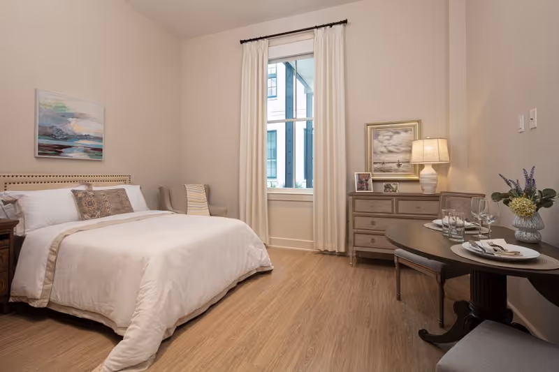 Bright, neatly furnished bedroom with a bed, dresser, window and small dining table.