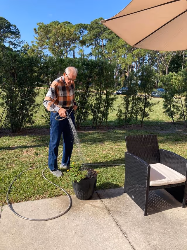 An elderly man wearing a plaid shirt and blue pants is watering a potted plant on a grassy lawn next to a concrete patio. There is a black wicker chair with a white cushion and a large beige patio umbrella nearby. Trees and parked cars are visible in the background under a clear blue sky.