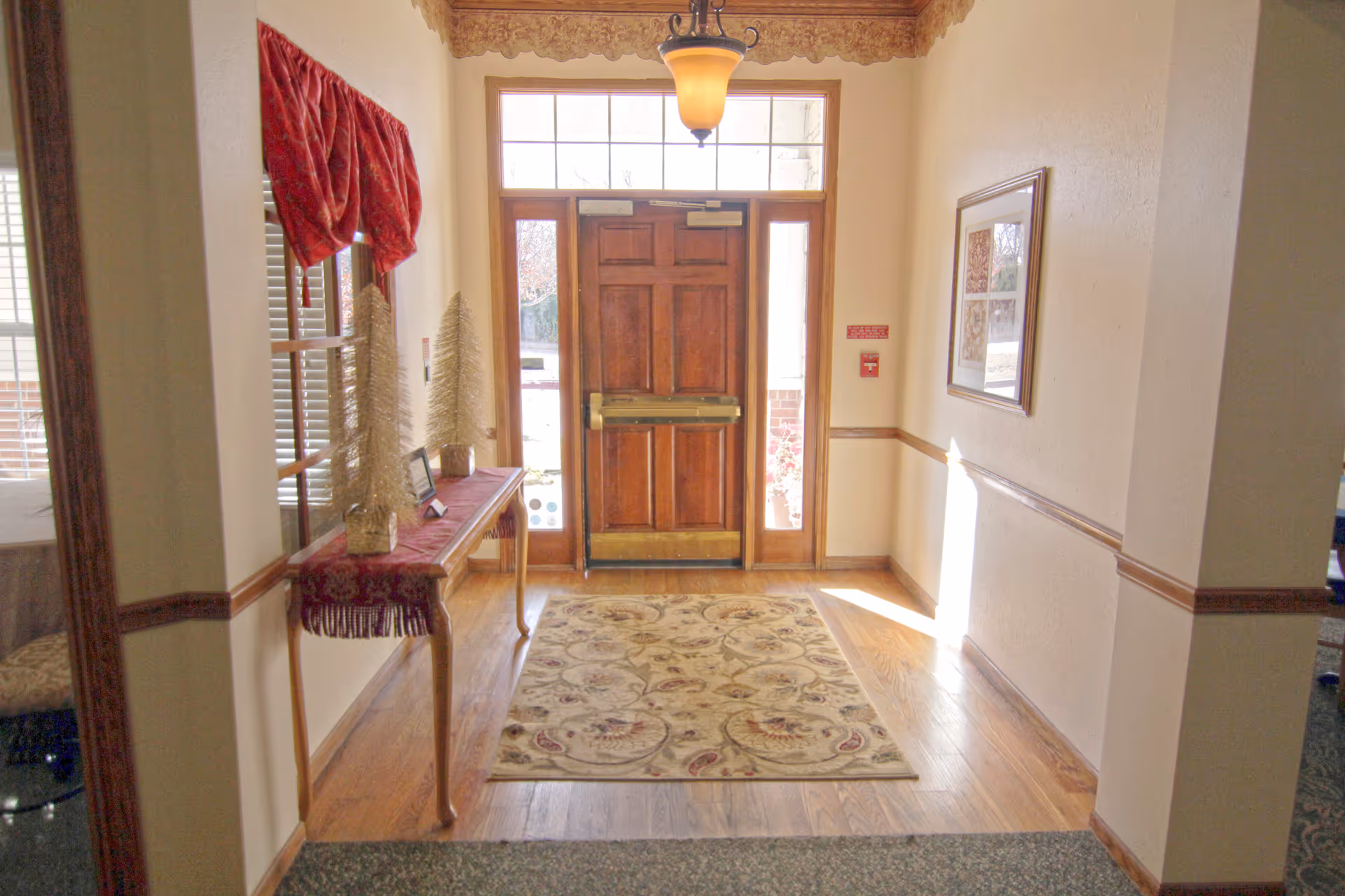 Interior view of an entrance hallway with a wooden door featuring glass panels on the sides and above. A decorative rug lies on the wooden floor in front of the door. To the left, a narrow wooden table with a red runner holds two decorative white Christmas tree figurines. A window with red curtains is above the table. The walls are light-colored with wooden trim, and a framed picture hangs on the right wall. A ceiling light fixture is centered above the door.