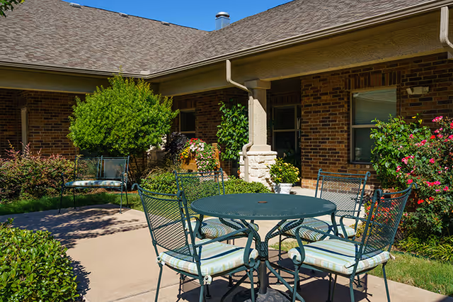 Outdoor patio area at Lynridge of Murphy Assisted Living & Memory Care with a round metal table and four chairs with cushions, surrounded by green bushes, flowering plants, and a brick building under a clear blue sky.