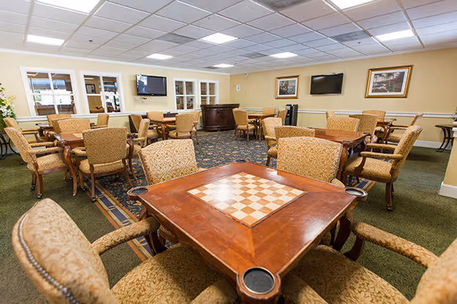 Communal activity room with upholstered chairs and game tables, a chessboard table in the foreground and wall-mounted TVs.