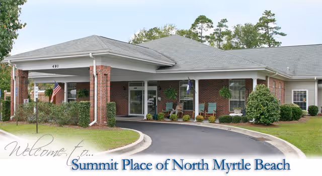 Front exterior view of a single-story brick building with a covered entrance, surrounded by greenery and a curved driveway. The building has a gray roof and several windows with white trim. There are American flags near the entrance and some outdoor seating visible.