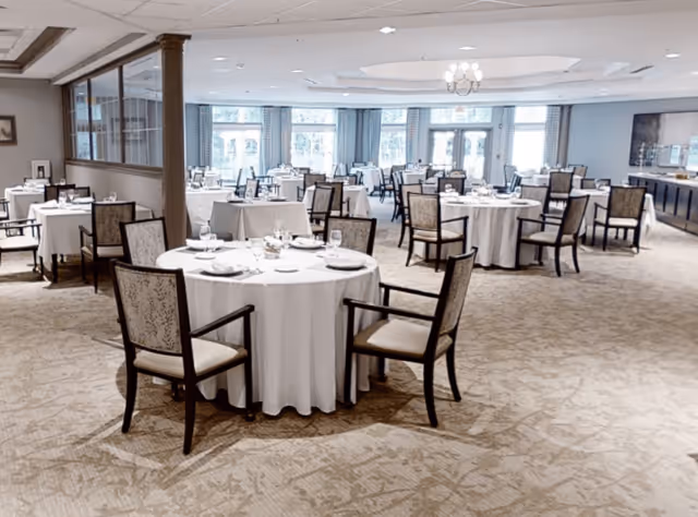 A spacious dining room with multiple round tables covered with white tablecloths, each set with plates, glasses, and silverware. The room has large windows with curtains allowing natural light to fill the space, and upholstered chairs arranged around the tables. The carpeted floor and ceiling lights create a warm and inviting atmosphere.