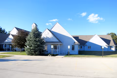 Exterior view of a single-story building with white siding and a gray roof under a clear blue sky. There are several trees and a small porch with a chair in front of the building. The area in front has a paved parking lot with handicap parking signs.