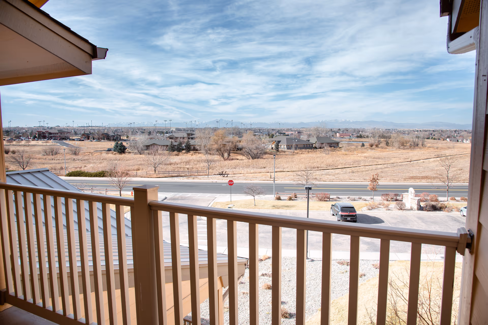 View from a balcony with beige railing overlooking a parking lot, a road, dry grassy fields, scattered trees, and distant mountains under a partly cloudy sky.