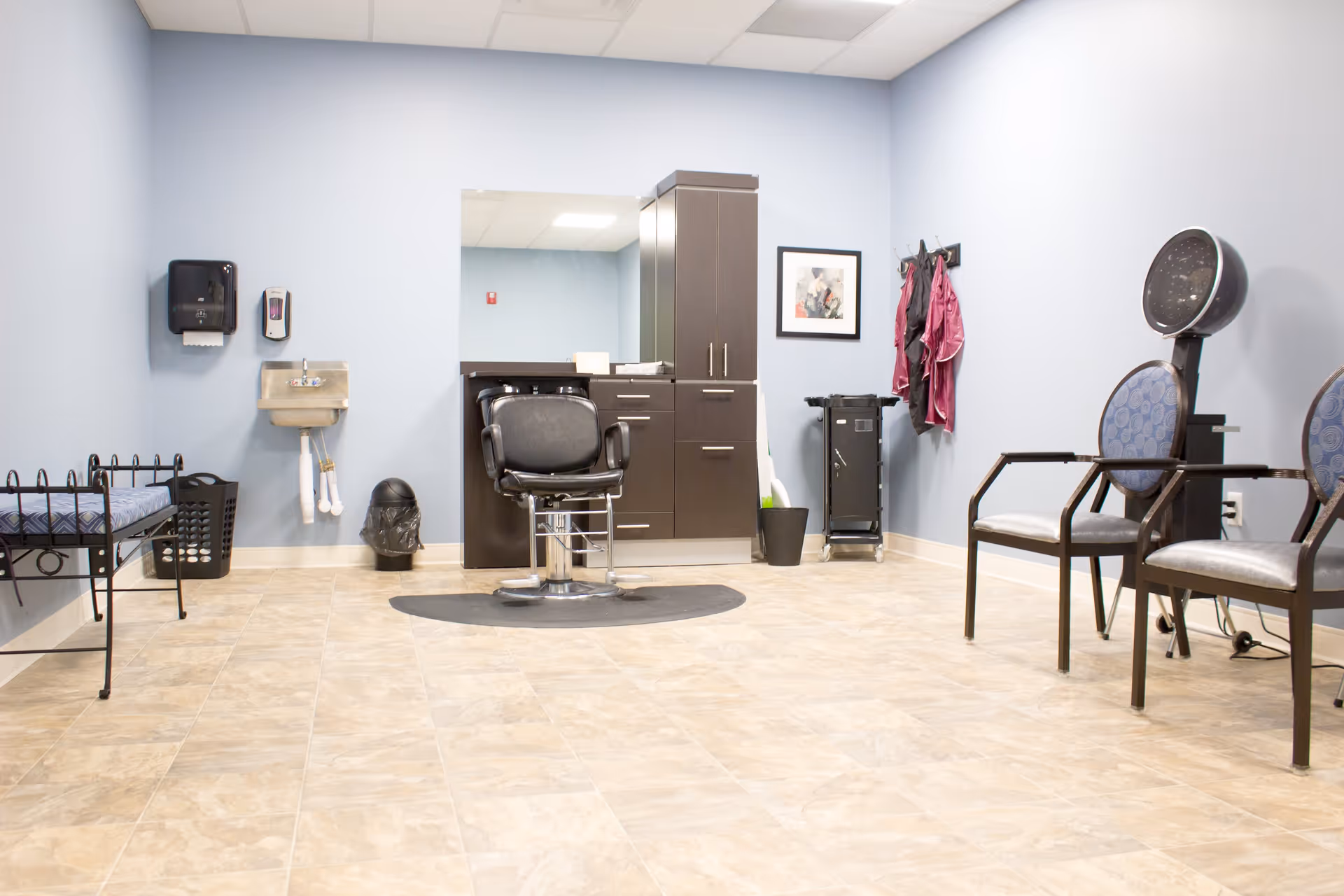 A clean and well-lit salon room with light blue walls and beige tiled floor. The room features a black salon chair in front of a large mirror mounted on a dark brown cabinet with drawers and cupboards. To the right, there are two chairs with blue patterned cushions and a black hair dryer hood. On the left side, there is a small metal bench with a blue cushion, a black laundry basket, a wall-mounted paper towel dispenser, a hand sanitizer dispenser, and a small stainless steel sink. A coat rack with hanging pink salon capes and a framed picture are visible on the right wall.