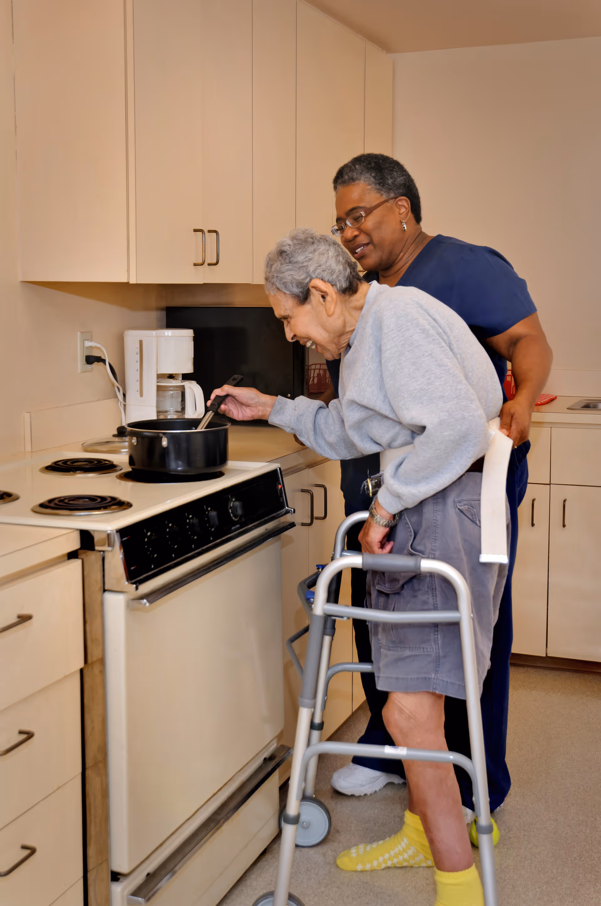 An elderly person using a walker is stirring a pot on a stove in a kitchen while being assisted by a caregiver standing behind them.