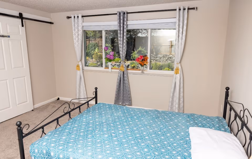 A bedroom with a black metal bed frame and a blue patterned bedspread. There is a window with three panels behind the bed, adorned with three curtains in white and gray colors tied with gold tassels. Outside the window, a garden with greenery and flowers is visible. To the left, there is a white sliding door with a sign that reads 'Restrooms'.