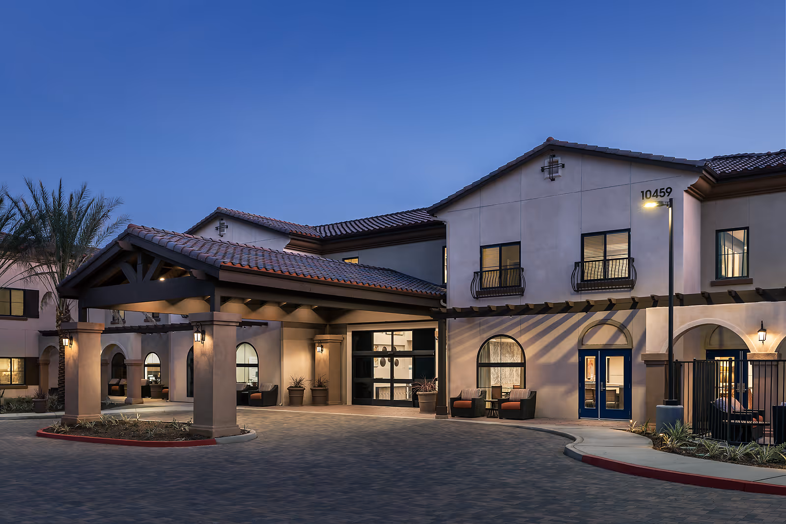 Exterior view of Cadence at Rancho Cucamonga senior living facility at dusk, showing a covered entrance with warm lighting, arched windows, and seating areas near the entrance.