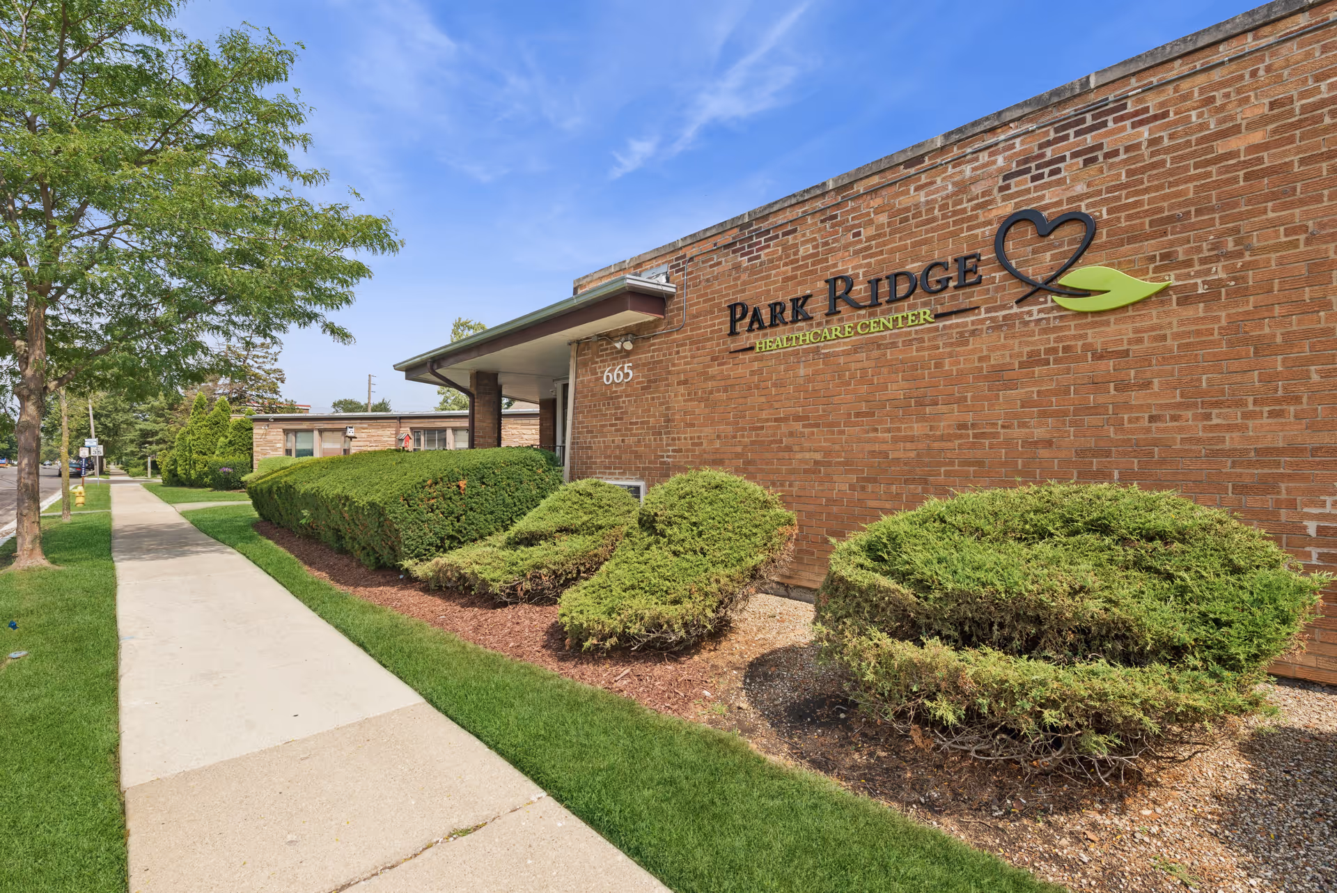 Brick exterior of Park Ridge Healthcare Center with its sign, landscaped shrubs and a sidewalk leading past the entrance.