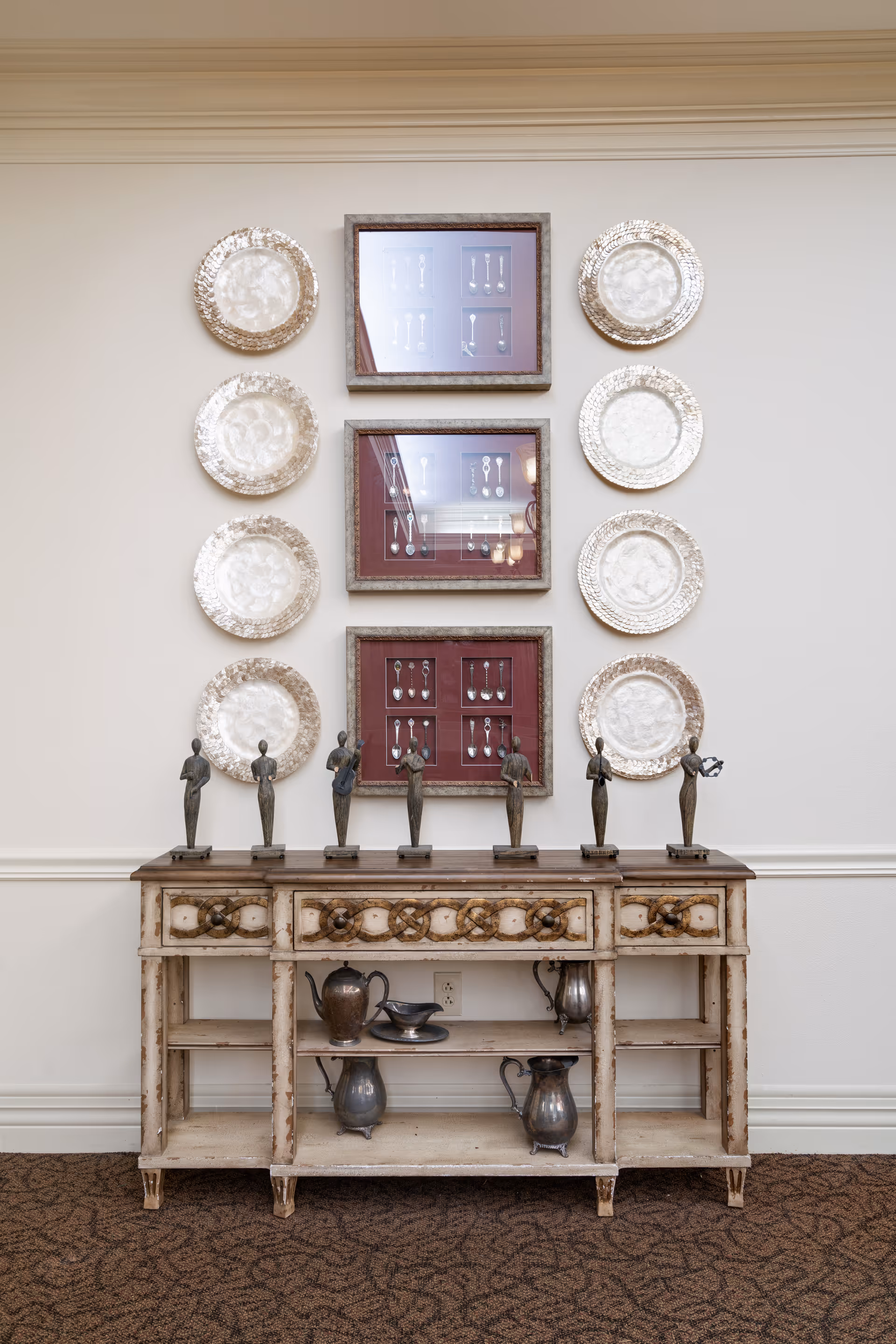 A decorative wooden sideboard with a distressed finish, displaying six small bronze statues on top. Above the sideboard, there are three framed displays of vintage silver spoons arranged vertically, flanked by six decorative silver plates mounted on the wall, three on each side. The setting is indoors with a beige wall and patterned carpeted floor.