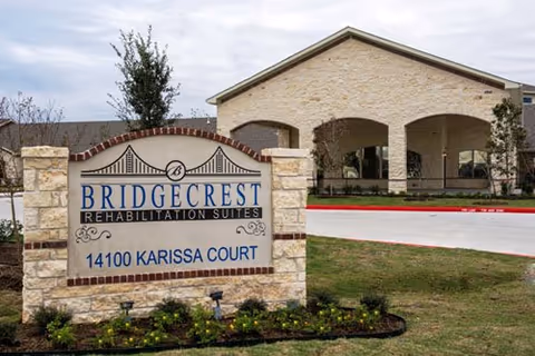 Stone sign for Bridgecrest Rehabilitation Suites at 14100 Karissa Court with a building featuring stone arches and a driveway in the background.