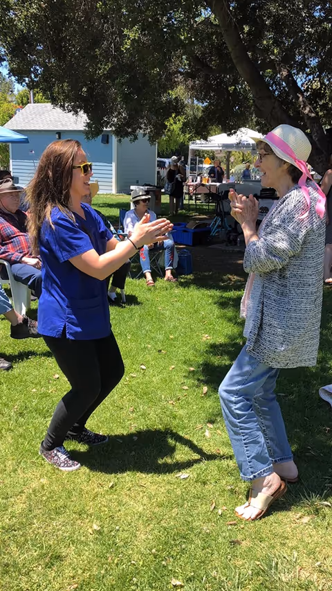 A woman in blue scrubs and sunglasses clapping hands and smiling with an elderly woman wearing a hat and glasses outdoors on a grassy area with other seated people and tents in the background.