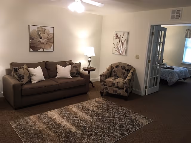 A cozy living room with a brown sofa adorned with white and patterned cushions, a matching patterned armchair, a small round wooden side table with a lamp, two framed floral artworks on the wall, and a large area rug covering the carpeted floor. A window with blinds and a glass-paneled door are visible in the background.