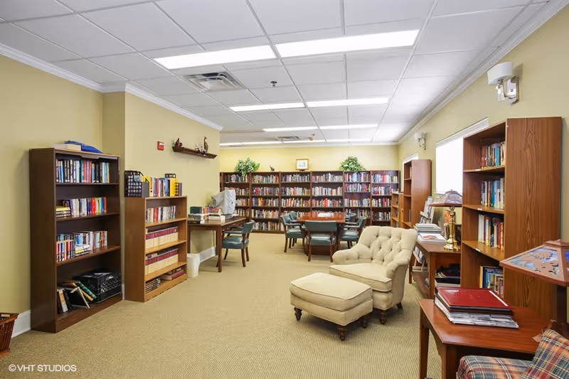 A well-lit library room with multiple wooden bookshelves filled with books along the walls. There are several tables with green cushioned chairs arranged for reading or studying. A beige upholstered armchair with a matching ottoman is positioned near a wooden side table with books and a lamp. The walls are painted light yellow, and the ceiling has recessed lighting panels.
