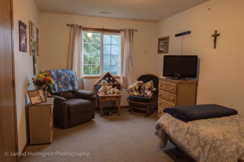 Cozy senior living bedroom with a bed, recliner, dresser and TV, a window with curtains, and stuffed animals on chairs.