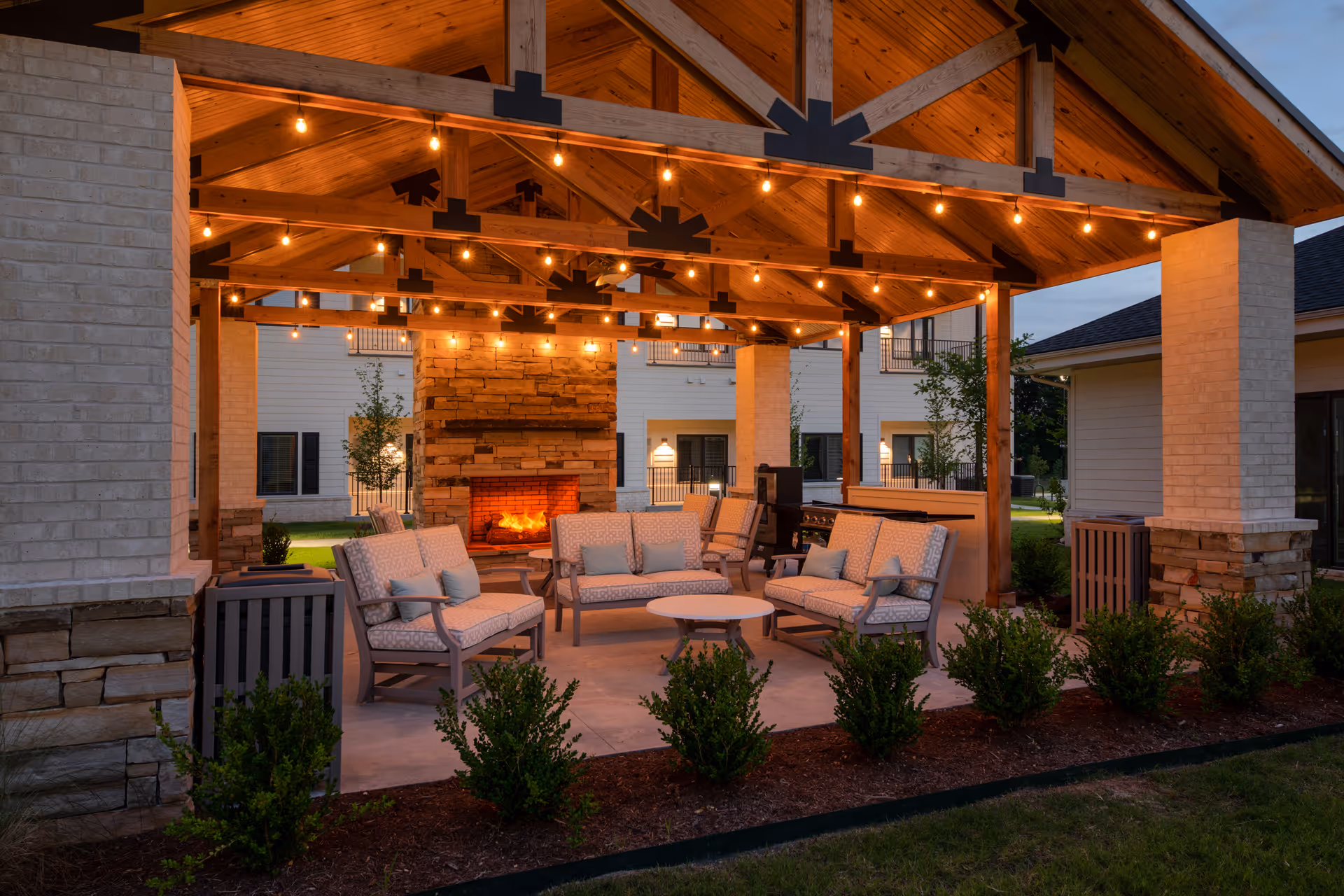 Outdoor covered seating area at dusk with cushioned chairs and sofas arranged around a round table in front of a stone fireplace. The wooden ceiling has exposed beams and string lights hanging, creating a warm and inviting atmosphere. Surrounding the area are small bushes and the exterior of the facility buildings.