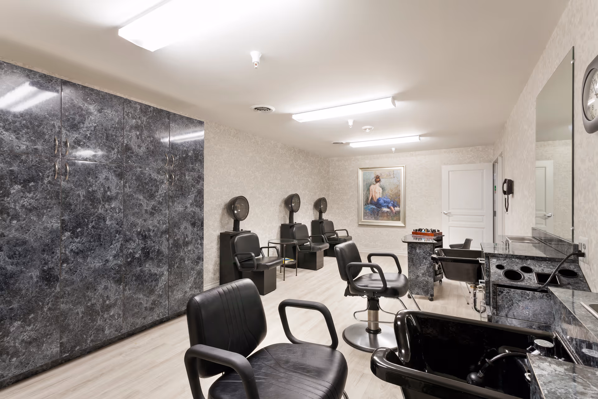 Interior view of a salon room with black salon chairs, hair dryers, a large mirror, and marble-patterned cabinets. The room has light-colored walls and flooring, with a framed painting on the far wall.