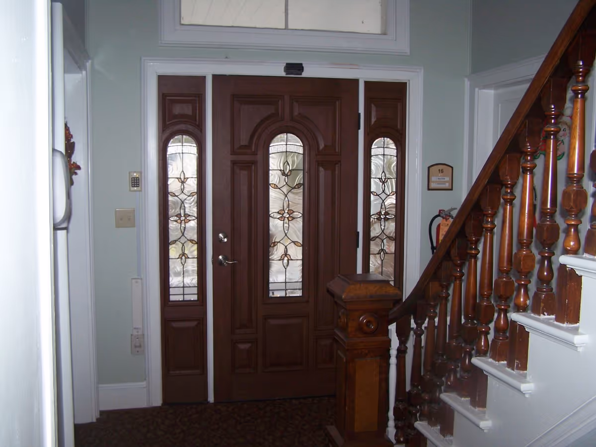 Interior view of an entrance area featuring a wooden front door with decorative glass panels on the door and sidelights. To the right, there is a wooden staircase with turned balusters and a newel post. The walls are painted light green, and there is a small plaque with the number 16 and a fire extinguisher mounted on the wall near the stairs.