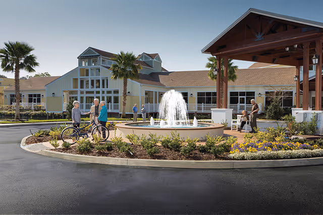Front entrance of a senior living facility with a central fountain, landscaped roundabout, palm trees, and residents chatting and walking nearby.