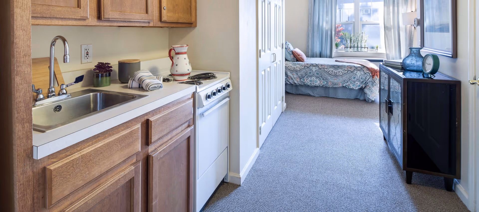 View of a small kitchen area with wooden cabinets, a stainless steel sink, and a white stove. Beyond the kitchen is a bedroom with a bed covered in a patterned bedspread, a window with blue curtains, and a dark wooden cabinet with decorative items on top.