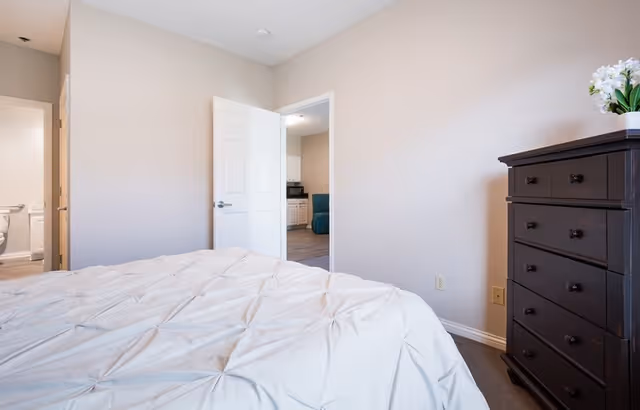A bedroom with a white textured bedspread on the bed, a dark wooden dresser with a small potted plant on top, and an open door leading to a bathroom and another room with white cabinets and a blue chair.