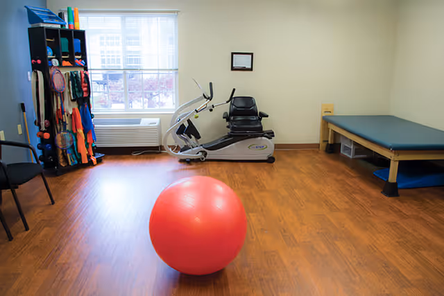 A small exercise room with a wooden floor, a large red exercise ball in the center, a recumbent exercise bike near a window with blinds, a black chair, a rack holding various exercise equipment including resistance bands and balls, and a padded treatment table with storage bins underneath.