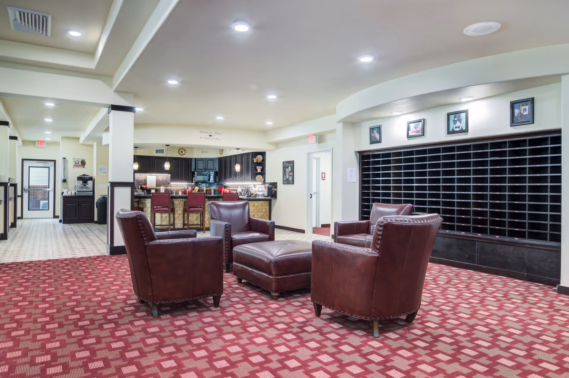 A spacious interior common area with four brown leather armchairs arranged around a matching ottoman on a red patterned carpet. In the background, there is a kitchen area with a counter and three red bar stools. The walls are light-colored with dark trim, and there are several framed pictures above a large black cubby storage unit. The ceiling has recessed lighting and a tray ceiling design.