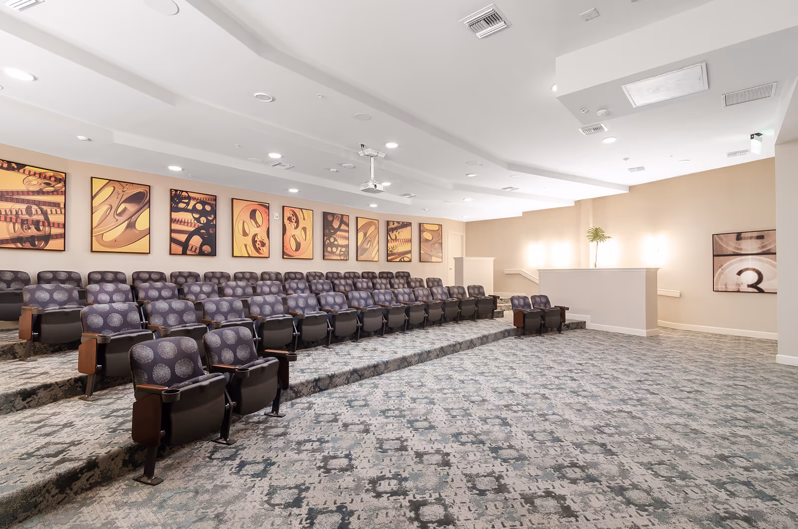 A small theater room with tiered seating featuring multiple rows of cushioned chairs with armrests and cup holders. The walls are decorated with framed artwork depicting film reels and movie-related imagery. The room has a patterned carpet and a white ceiling with recessed lighting and a projector mounted in the center.