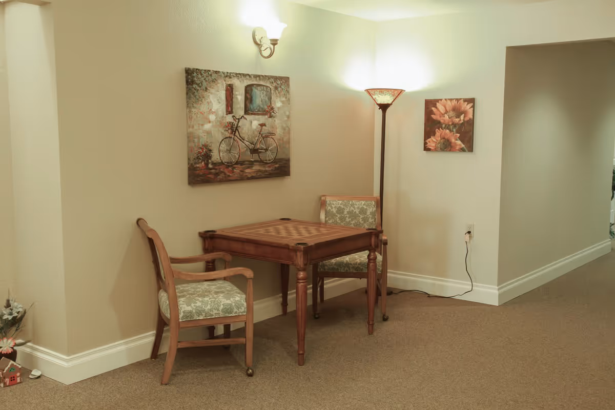 A small corner seating area with a wooden table featuring a checkerboard pattern and two wooden chairs with floral cushions. The walls are beige and decorated with two paintings, one of a bicycle and the other of sunflowers. A wall sconce and a floor lamp provide soft lighting. The floor is carpeted, and there is an electrical outlet with a plugged-in cord on the wall.