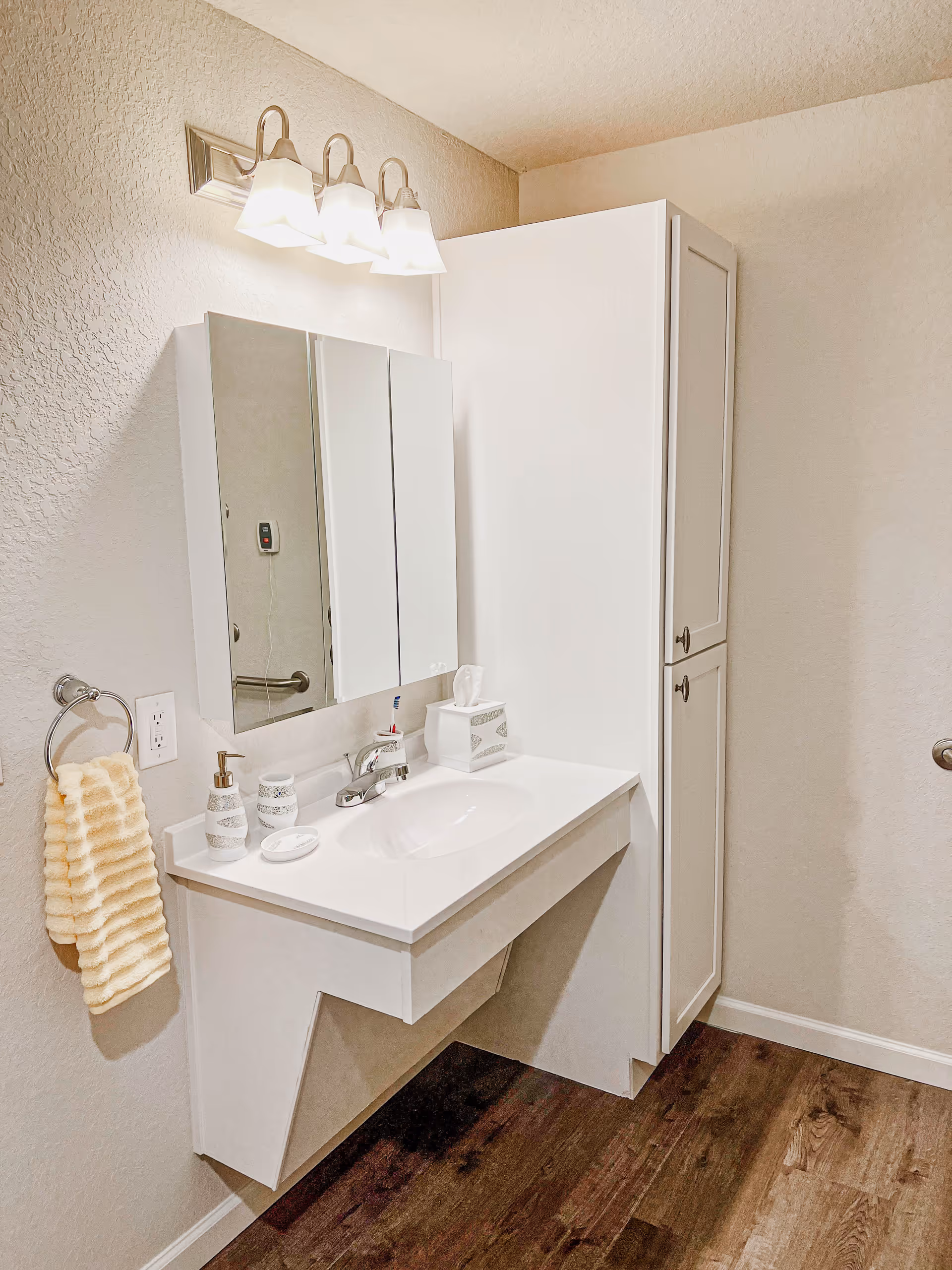 A clean and modern bathroom vanity area with a white sink, a wall-mounted mirror cabinet, a three-light fixture above the mirror, a beige textured hand towel hanging on a ring, and a tall white storage cabinet next to the sink. The floor is dark wood, and the walls are light beige.