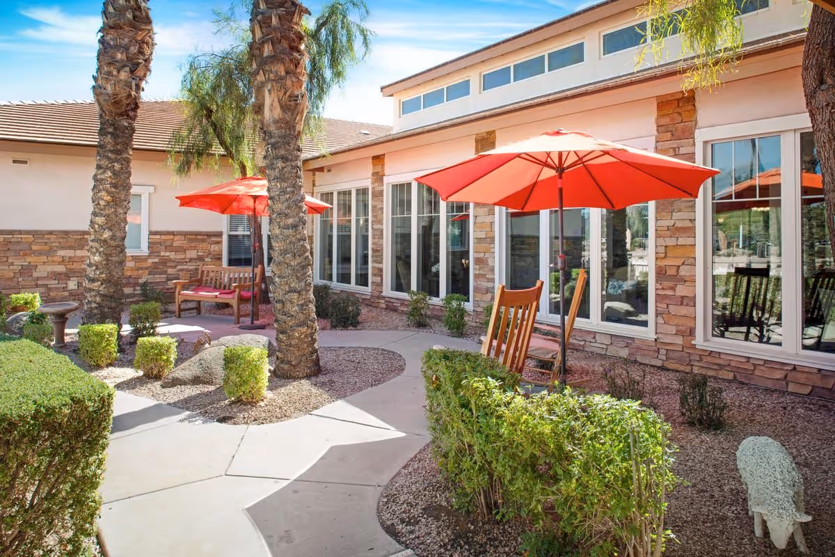 Sunny courtyard with palm trees, red patio umbrellas and wooden seating beside a stone-faced building with large windows.