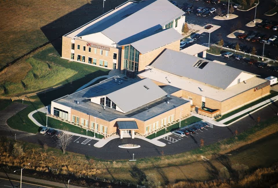 Aerial view of a large multi-wing brick building with multiple parking areas and surrounding green lawns, likely a senior living or healthcare facility.